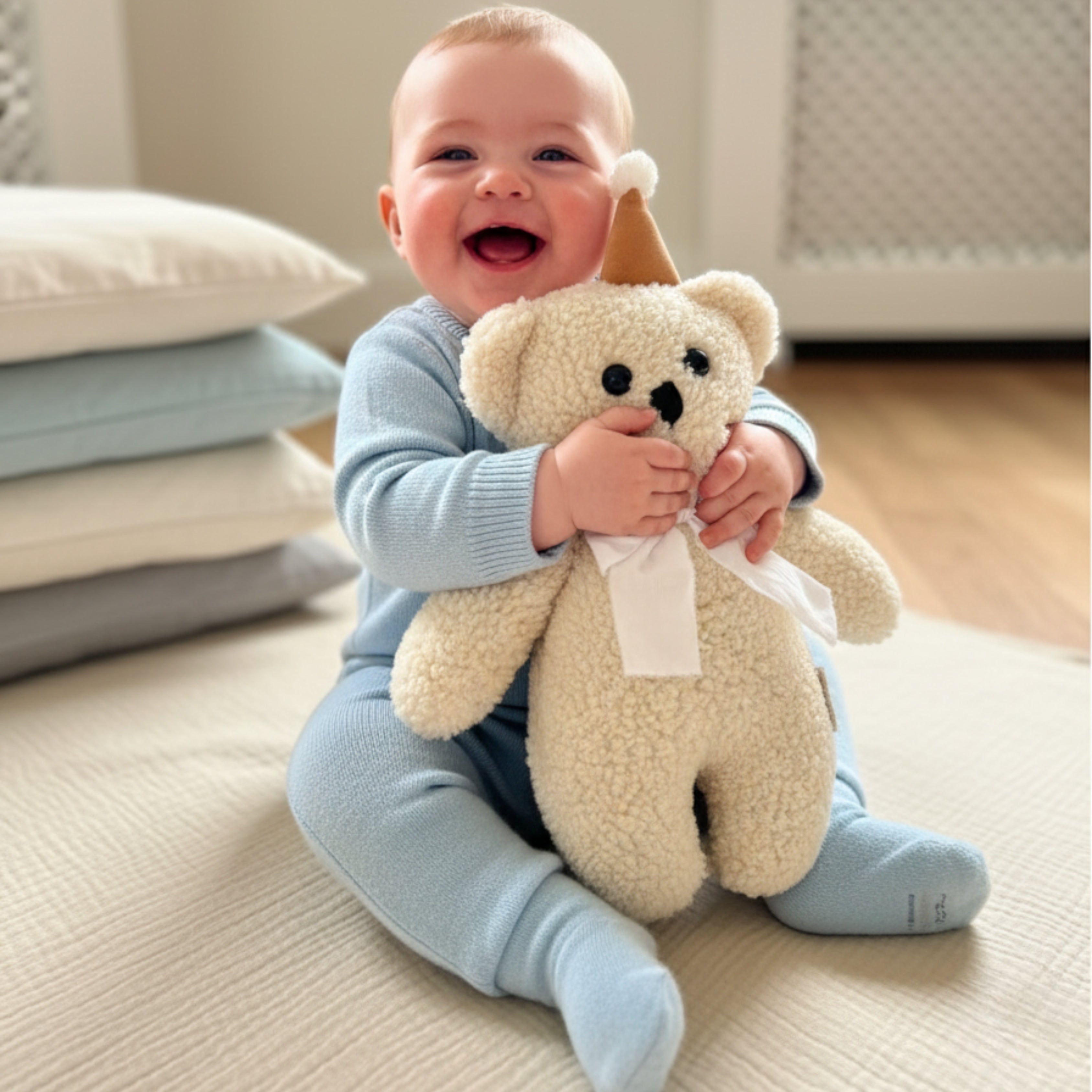 A smiling baby sitting on a soft mat while holding Honey the Bear Sleep Companion — an organic plush that brings comfort, joy, and emotional security.