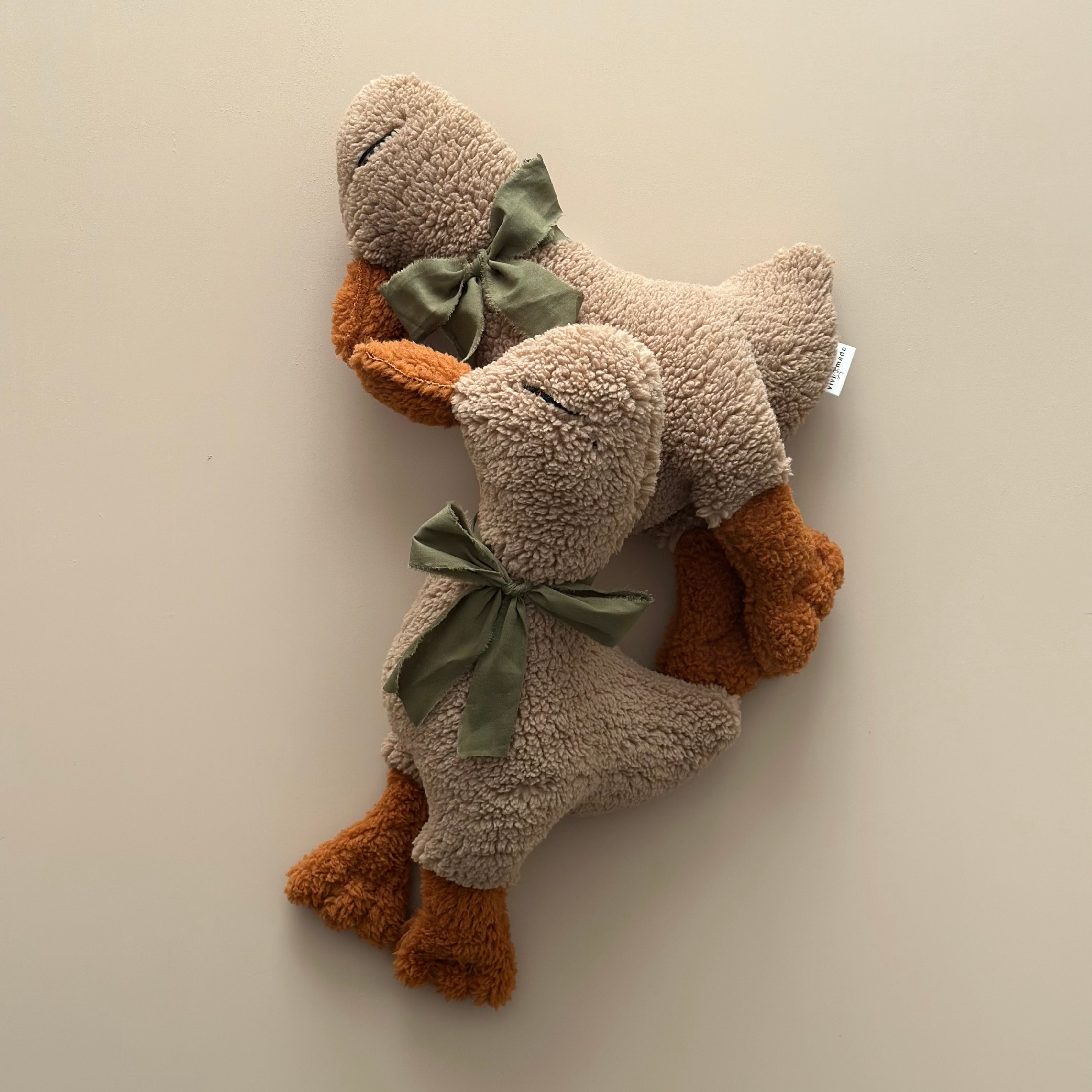 Two handmade Luna the Duck Sleep Companions resting together on a neutral beige surface, featuring soft brown tones and green fabric bows.