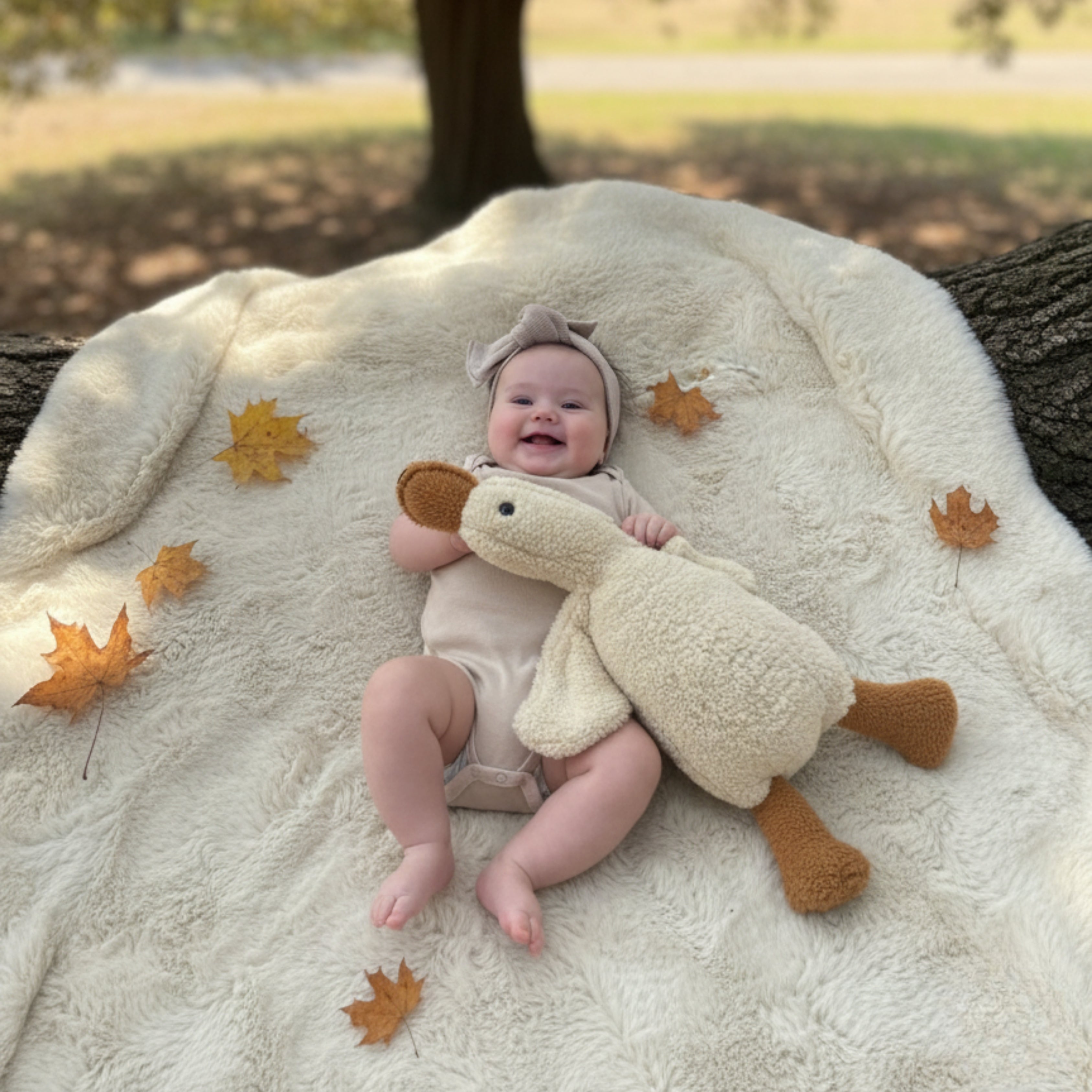 A smiling baby lying on a soft blanket surrounded by autumn leaves, cuddling Ollie the Duck Sleep Companion — a handcrafted plush designed for comfort and calm.