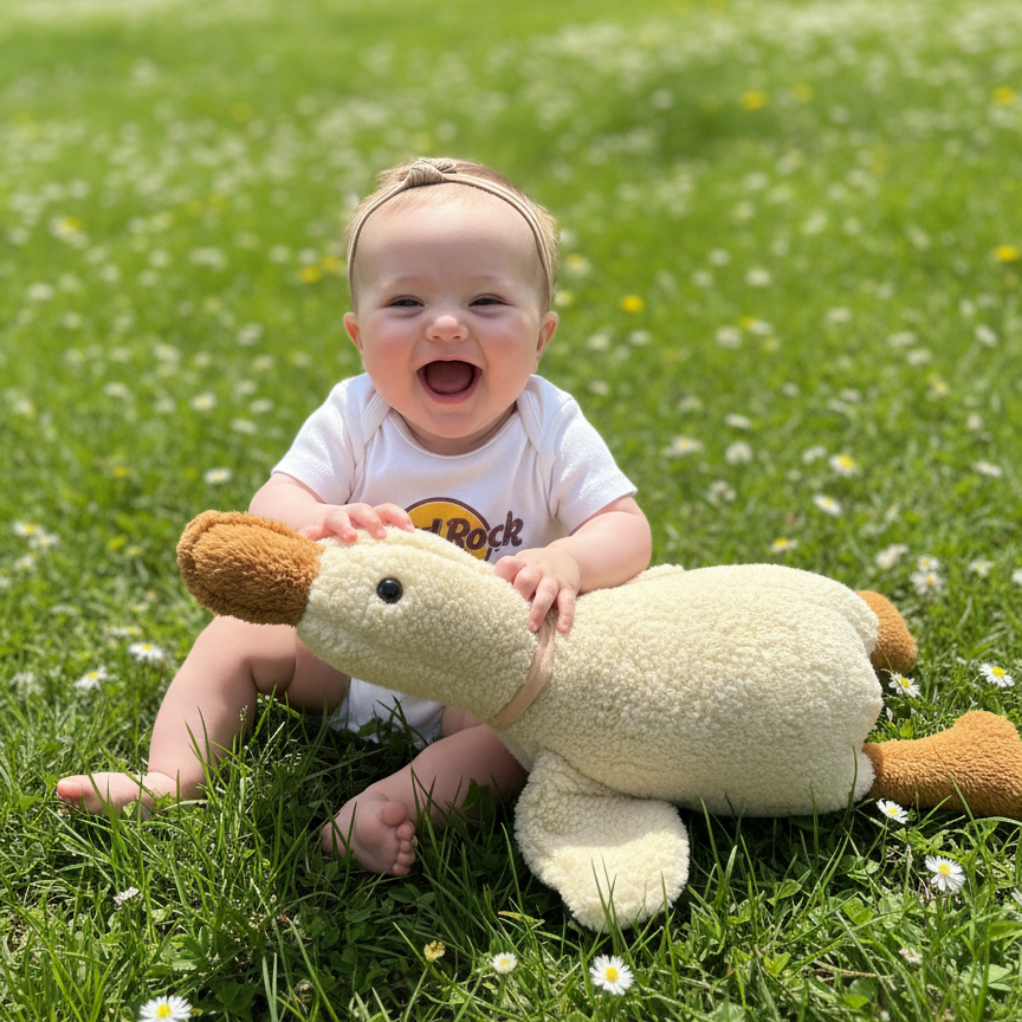 A cheerful baby sitting among daisies, laughing while holding Ollie the Duck Sleep Companion — a handmade plush duck that brings calm joy to spring playtime.